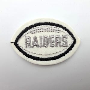 May include: An oval-shaped embroidered patch featuring a white felt base with a black stitched border. The center displays the word RAIDERS in silver thread, stylized to resemble a football with silver stitching details at the top and bottom.