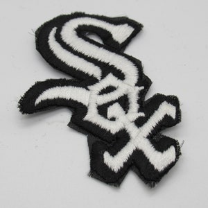 May include: A black and white embroidered patch featuring the Chicago White Sox logo. The design consists of a stylized, interlocking S and O letter combination with a thick black border and white thread detailing throughout the emblem.