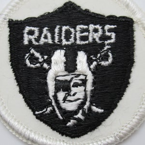 May include: A circular embroidered patch featuring a black shield shape on a white background. The shield contains the white text RAIDERS above a white and black embroidered graphic of a football player wearing a helmet and an eye patch.