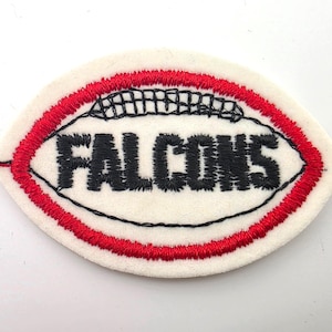 May include: An oval-shaped embroidered patch featuring a white background with a red stitched border. The center displays the word FALCONS in bold black thread, with a black stitched football lace design positioned directly above the text.