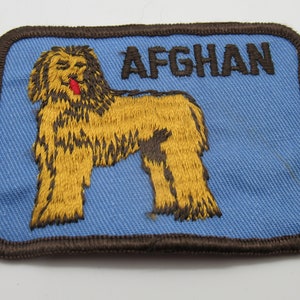 May include: A rectangular embroidered patch with a light blue background and a dark brown stitched border. The patch features a golden-brown Afghan Hound dog with a small red tongue and the word AFGHAN embroidered in dark brown capital letters.
