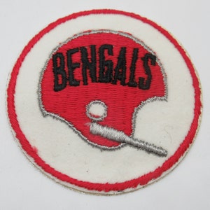 May include: A circular embroidered patch featuring a red football helmet with the word BENGALS stitched in black letters across the top. The helmet includes a silver facemask and is set against a white background with a red stitched border.