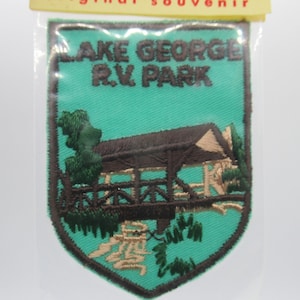 May include: A shield-shaped embroidered patch with a teal background and a dark brown border. The text reads LAKE GEORGE R.V. PARK in dark brown letters. The design features a brown covered bridge, green trees, and tan water elements.