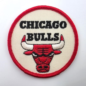 May include: A circular embroidered patch featuring the Chicago Bulls logo. The design includes the text CHICAGO BULLS in black thread above a red bull head with white horns, all set against a cream background with a thick red stitched border.