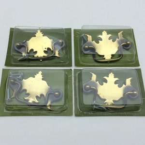 May include: Four brass-colored metal drawer pulls with ornate backplates and swinging handles, each individually sealed in clear plastic blister packaging on green cardboard backing, accompanied by small silver-colored metal mounting screws.