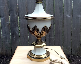 May include: A white and gold table lamp with a decorative base and a white shade. The lamp is sitting on a wooden table.