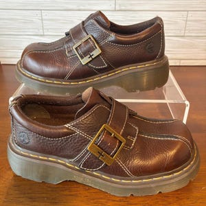 May include: A pair of brown leather shoes with a buckle closure and white stitching. The shoes have a platform sole and a classic design. The shoes are approximately 10 inches long.