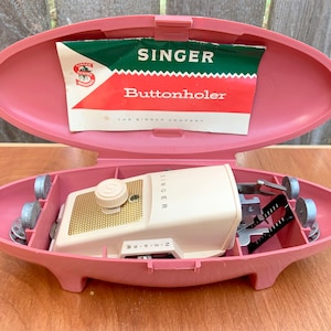 May include: A vintage pink Singer buttonholer in its original case. The buttonholer is white with gold accents and has a knob on the top. The case is oval-shaped and has a hinged lid. The case also includes a small instruction booklet that reads "Singer Buttonholer The Singer Company."