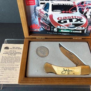 May include: A wooden box containing a John Force 7 Time Funny Car Champion Serialized Limited Edition Knife, a silver coin, and a card with details about the knife. The box is open and the knife is visible. The knife has a bone handle and a gold blade.