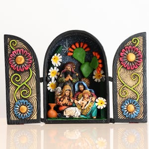 May include: A colorful, hand-painted nativity scene displayed in a three-panel wooden frame. The central panel features figures of Mary, Joseph, and baby Jesus, surrounded by flowers and cacti. The side panels are decorated with large, stylized flowers.