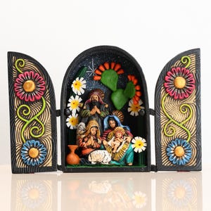 May include: A colorful, hand-painted nativity scene displayed in a three-panel wooden frame. The central panel features figures of Mary, Joseph, and baby Jesus, surrounded by flowers and cacti. The side panels are decorated with large, stylized flowers.