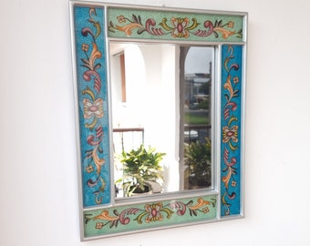 May include: A silver framed mirror with a blue and green painted border featuring floral designs. The mirror has a clear reflection of a plant and a window.