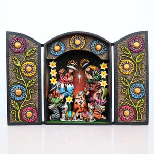 May include: A carved wooden nativity scene with a central arched section and two side panels. The scene depicts figures in colorful clothing, with floral and vine designs on the panels. The color palette includes black, brown, yellow, blue, pink, and green.