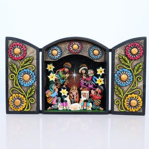 May include: A three-panel, hand-painted nativity scene. The black frame is adorned with colorful floral designs. The central panel depicts the holy family and onlookers. The figures are dressed in vibrant traditional clothing.