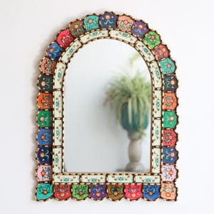 May include: An arched mirror with a decorative, scalloped frame. The frame is a mosaic of small, hand-painted tiles in various colours, including red, blue, green, and orange. The mirror has a gold-coloured trim.