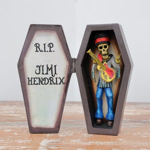 May include: A wooden coffin with the words "R.I.P. Jimi Hendrix" on the front. The coffin is open and a skeleton figure with a guitar is inside. The skeleton is wearing a blue shirt, blue jeans, and a red headband.