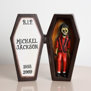 May include: A decorative coffin with the text "R.I.P. MICHAEL JACKSON 1958-2009". Inside is a skeleton figure wearing a red jacket and pants. The coffin is brown with a white interior.