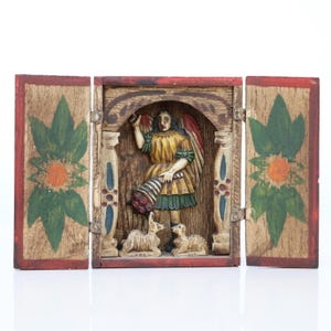 May include: A wooden, hand-carved triptych featuring a central angel figure with a horn and two small animals at the base. The angel wears a yellow robe and has red and white wings. The side panels have painted floral designs with red borders.