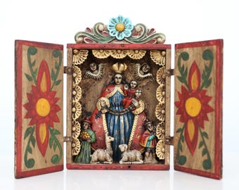 May include: A wooden religious diorama with a central carved figure in a red and blue robe, holding a child. The diorama has two opening doors with floral designs and a decorative top with a blue flower. The interior features angels and figures.