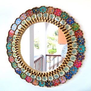Op de afbeelding: Een ronde spiegel met een kleurrijk, mozaïekachtig frame. Het frame is gemaakt van kleine, individuele tegels in verschillende tinten blauw, groen, roze, geel en paars. De tegels zijn in een cirkelvormige patroon rond de spiegel gerangschikt.