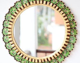 May include: A round mirror with a gold frame and a green and white floral design. The mirror is surrounded by a scalloped edge.