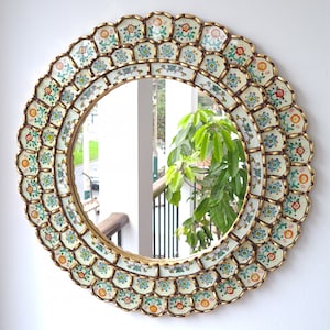 Puede incluir: Un espejo redondo con un marco decorativo hecho de azulejos blancos con detalles dorados y diseños florales coloridos. El espejo refleja una vista de una planta verde y un balcón.