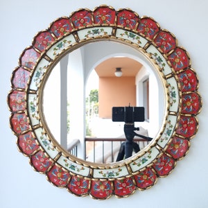 Puede incluir: Un espejo redondo con un marco decorativo y ornamentado. El marco está hecho de madera y está pintado con acentos rojos, blancos y dorados. El marco presenta un borde festoneado y diseños florales intrincados.