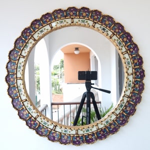 May include: A round mirror with a gold frame decorated with purple and white floral designs. The mirror is reflecting a tripod with a camera on top.
