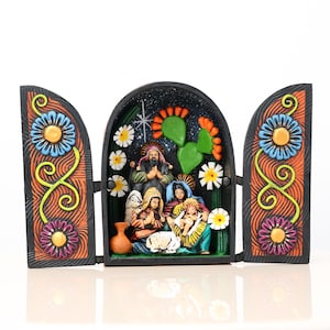 May include: A hand-painted, wooden nativity scene with a black background and colorful floral and cactus accents. The scene depicts the holy family, angels, and a lamb. The doors are decorated with blue, yellow, and purple flowers.