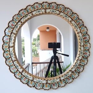 Puede incluir: Un espejo decorativo redondo con borde festoneado. El marco es dorado con un patrón floral en tonos rojos, azules y verdes. El espejo refleja un balcón y un trípode.