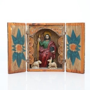 May include: A wooden religious diorama featuring a carved figure in red and green robes, holding a staff and book, with a gold halo. Two lambs are at the figure's feet. The diorama has two hinged doors with blue and orange floral designs.