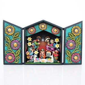 May include: A colorful, hand-carved nativity scene with a central structure and two hinged side panels. The scene features figures in traditional clothing, floral accents, and a star-filled backdrop. The frame is decorated with flowers and vines.