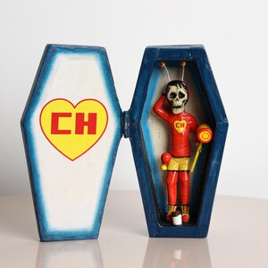 May include: A colorful, hand-painted figurine of a skeleton character in a coffin-shaped box. The skeleton wears a red shirt with a yellow heart and "CH" logo, yellow shorts, and red pants. The coffin is blue with a white interior.