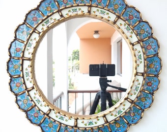 May include: A round mirror with a decorative frame. The frame is made of wood and is painted with blue, white, and gold paint. The frame has a scalloped edge and is decorated with floral designs. The mirror is reflecting a room with a white ceiling and a brown wall.