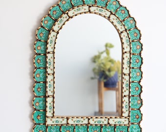 May include: An arched wall mirror with a decorative frame. The frame features a scalloped edge with turquoise and floral patterned tiles, accented with gold trim. The mirror reflects a room with a plant.