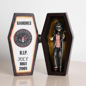 May include: A dark brown coffin-shaped box with the word "RAMONES" and "R.I.P. JOEY 1951 2001" printed on the lid. Inside, a figurine of a skeleton wearing a striped shirt, black jacket, and sunglasses, holding a microphone.