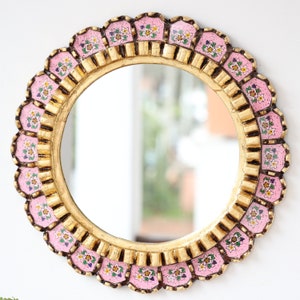 May include: A round mirror with a gold frame and pink and white floral details. The frame is ornate and has a scalloped edge.
