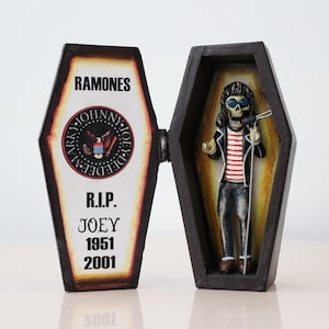 May include: A decorative coffin-shaped box with the word "RAMONES" at the top. The left side has a circular emblem and the text "R.I.P. JOEY 1951 2001". The right side contains a skeleton figure in a red and white striped shirt, holding a microphone.