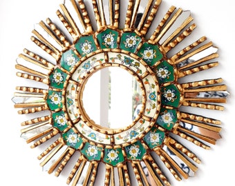 May include: A gold sunburst mirror with a green and white floral design. The mirror has a round shape and is surrounded by a decorative frame.