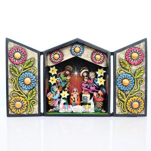 May include: A three-panel, hand-painted nativity scene. The black frame is decorated with colorful floral designs. The central panel depicts the holy family and surrounding figures. The figures are dressed in vibrant colors, and the scene is set against a starry backdrop.