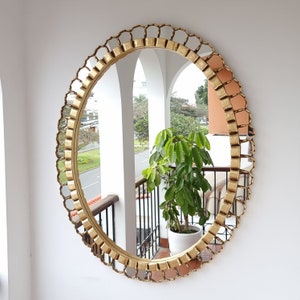 May include: A gold-toned round mirror with a decorative, ornate frame. The frame is made of metal and has a scalloped design. The mirror reflects a view of a balcony with plants and trees.