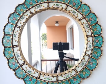 May include: A round mirror with a decorative frame featuring a repeating pattern of blue and white floral designs. The frame is made of gold metal and has a scalloped edge.