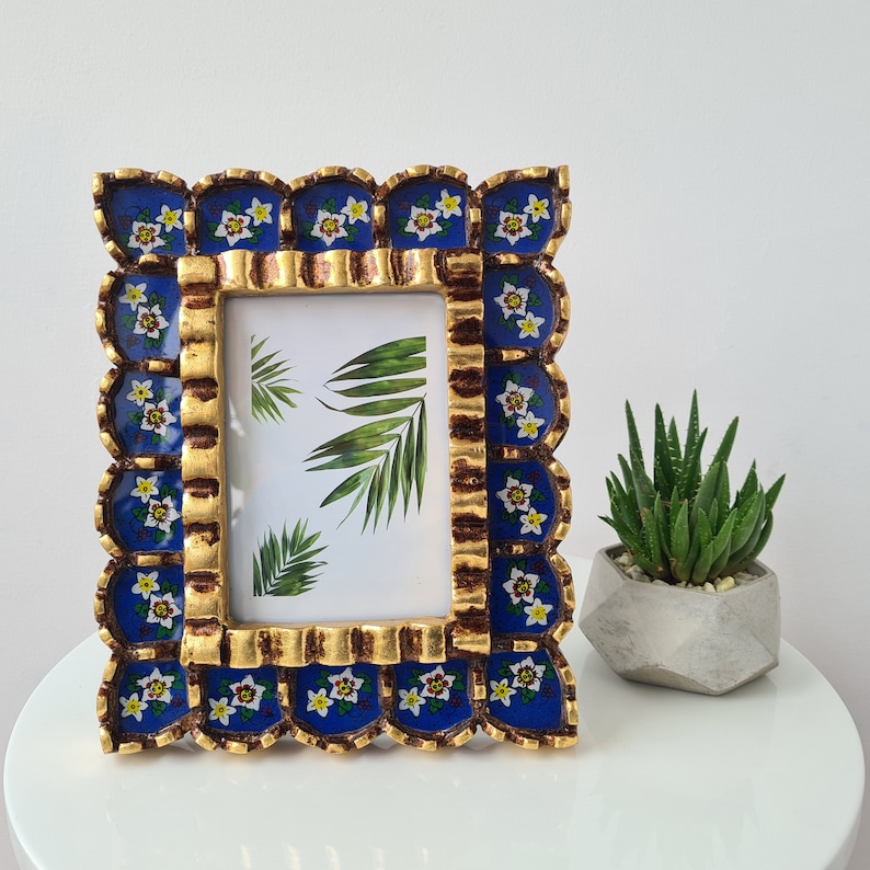 Pu&ograve; includere: Una cornice per foto blu e oro con un bordo a festoni e disegni floreali. La cornice ha un passepartout bianco e un vetro trasparente.