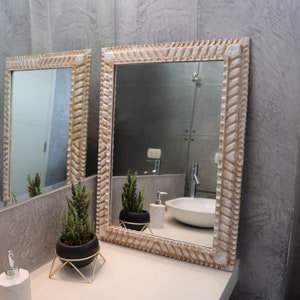 May include: A white bathroom mirror with a carved wooden frame. The mirror is hanging on a gray wall and reflects a bathroom sink, a plant, and a soap dispenser.