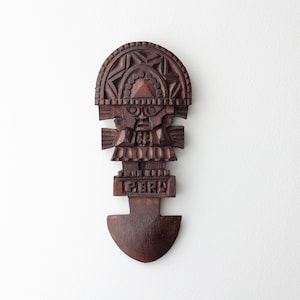 Puede incluir: Una talla de madera marrón oscuro de una figura con un tocado, que lleva una falda estampada y sostiene un bastón. La talla está etiquetada como "PERU".