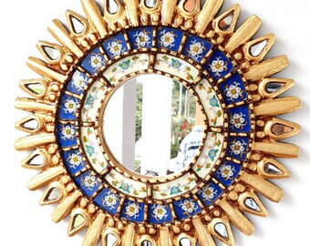 May include: A gold sunburst mirror with a blue and white tile design. The mirror has a round shape and is surrounded by a decorative frame.