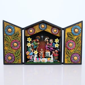 May include: A three-panel, hand-carved nativity scene. The black frame features colorful floral designs on the side panels. The central panel depicts the holy family and other figures. The figures are painted in bright colors, with gold accents.