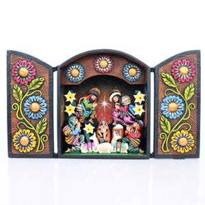 May include: A colorful, hand-painted nativity scene displayed in a wooden triptych. The artwork features figures in vibrant clothing, set against a starry backdrop. The frame is adorned with floral designs in shades of pink, blue, and yellow.