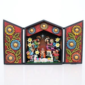 May include: A colorful, hand-painted nativity scene in a triptych design. The central scene depicts the holy family and the three wise men. The frame is black with red panels adorned with floral patterns in yellow, blue, and orange.