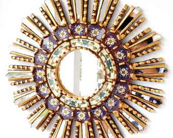 May include: A gold sunburst mirror with a round, purple and white floral design. The mirror has a decorative, ornate frame with a sunburst pattern.
